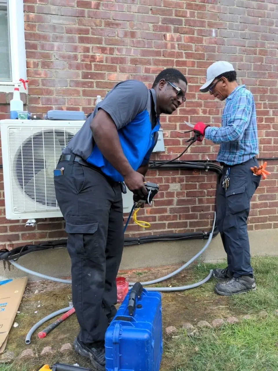 Two technicians at a mini-split outdoor unit, one holding a meter and one working on the wiring